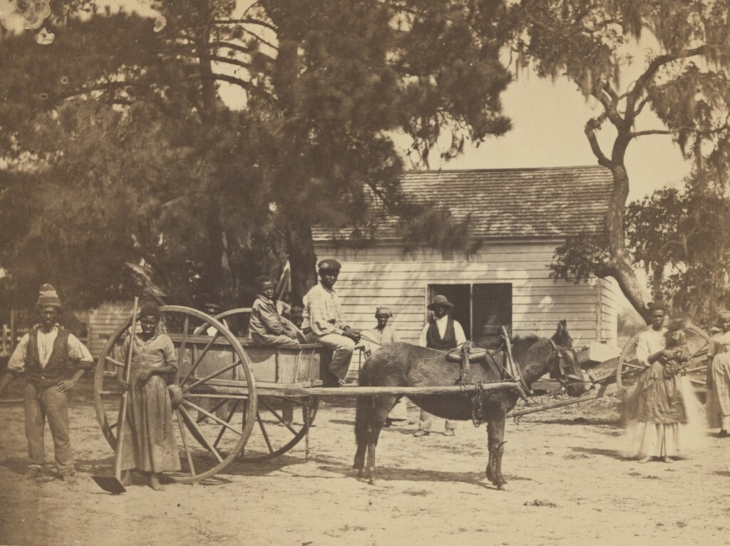 A posed photograph with 9 enslaved people, a horse cart, and a small building and trees.